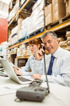 Manager working on laptop and talking on phone at desk in a large warehouse