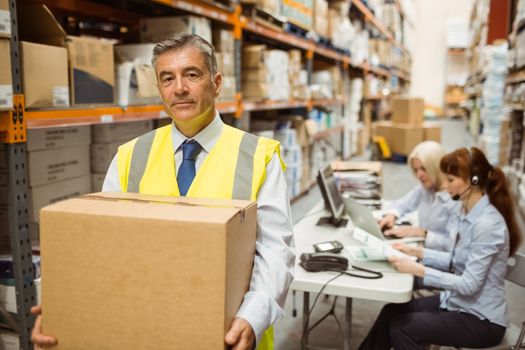 Warehouse manager smiling at camera carrying a box in a large warehouse
