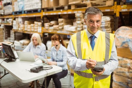 Smiling manager wearing yellow vest using handheld in a large warehouse