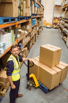 Worker with trolley of boxes smiling at camera in a large warehouse