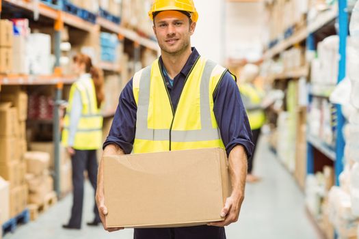 Warehouse worker smiling at camera carrying a box in a large warehouse