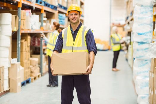 Warehouse worker smiling at camera carrying a box in a large warehouse