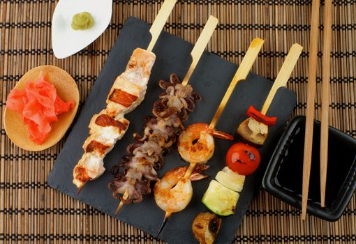 Grilled Salmon, Octopuses, Shrimps and Vegetables on Wooden Stick with Ginger, Soy Sauce and Wasabi on Straw Mat background. Top View