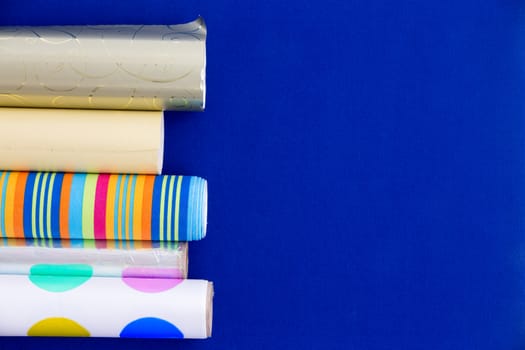 Colorful rolls of patterned and metallic wrapping paper arranged as a side border over a blue background with copyspace