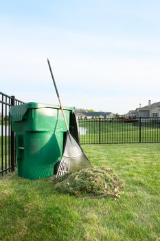 Yard maintenance in spring cleaning the lawn and raking up grass clippings after mowing with a pile of grass and rake leaning on a plastic bin for composting