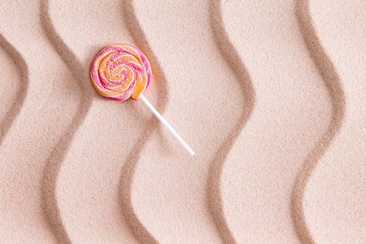 Nothing tastes crunchier than a sandy lollipop at the seaside on a summer vacation with a twirled round sucker on white beach sand with a decorative wavy pattern and copy space