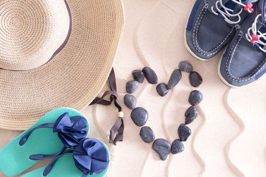 Young summer love at the beach in a conceptual image with a straw sunhat, slip slops and boys shoes surrounding a pebble heart on golden beach sand with a decorative wavy pattern