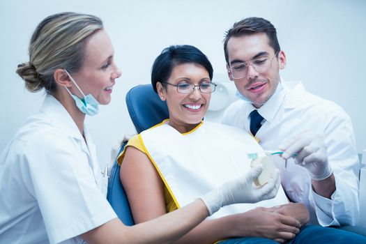 Male dentist with assistant showing woman how to brush teeth in the dentists chair