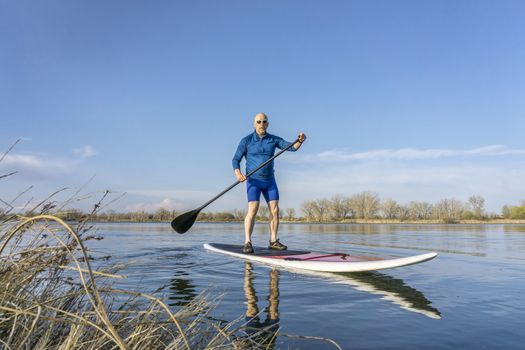 Senior male on stand up paddling (SUP) board. Early spring on calm lake in Colorado.