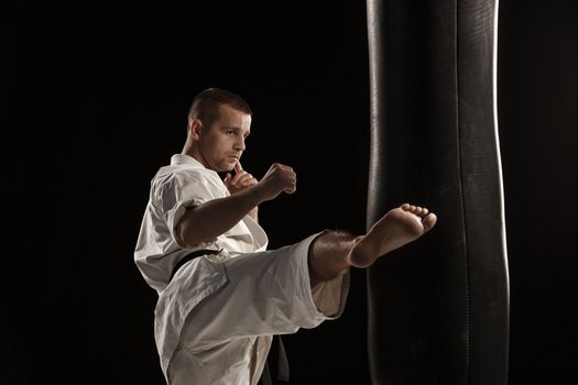 Man in white kimono training karate round kick in a punching bag over black background