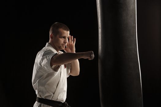 Man in white kimono training karate in a punching bag over black background