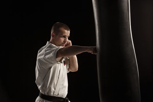 Man in white kimono training karate in a punching bag over black background