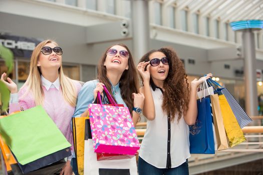 shopping, sale, happy people and tourism concept - three beautiful girls in sunglasses with shopping bags in mall