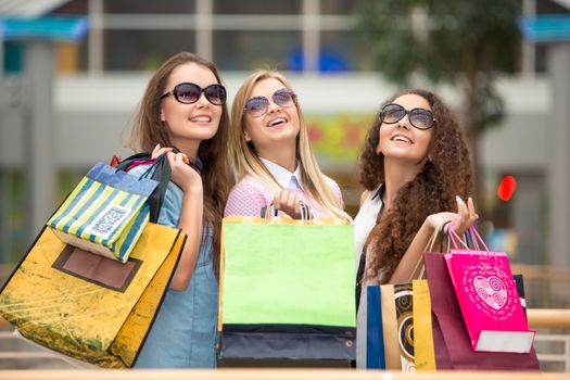 shopping, sale, happy people and tourism concept - three beautiful girls in sunglasses with shopping bags in mall