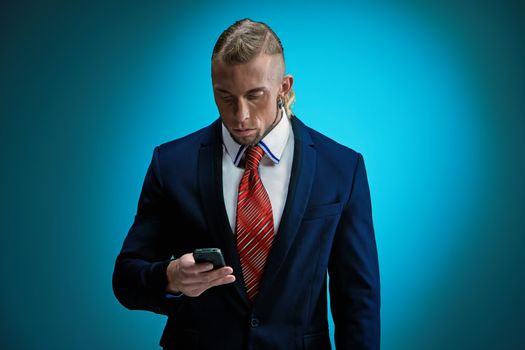 Portrait of an attractive businessman with phone wearing black suit. Blonde hair. On blue background