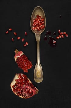 Delicious ripe pomegranate seeds with silver spoon on black background, top view. Healthy exotic fruit eating.