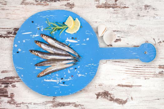 Delicious fresh sardines on wooden kitchen board with lemon, rosemary and colorful peppercorns on white textured wooden background. Culinary healthy cooking.