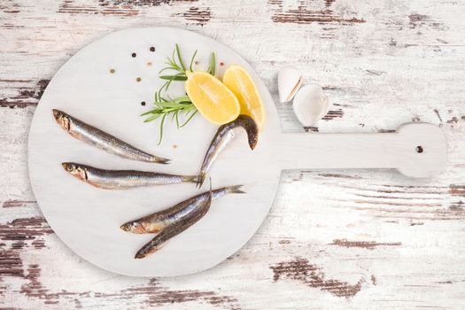 Delicious fresh sardines on wooden kitchen board with lemon, rosemary and colorful peppercorns on white textured wooden background. Culinary healthy cooking.