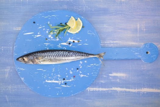 Delicious fresh mackerel fish on wooden kitchen board with lemon, rosemary and salt flakes on blue textured wooden background. Culinary healthy cooking.