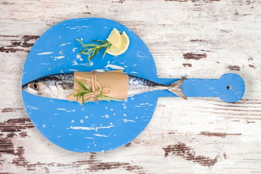 Delicious fresh mackerel fish on wooden kitchen board with lemon, rosemary and salt flakes on white textured wooden background. Culinary healthy cooking.