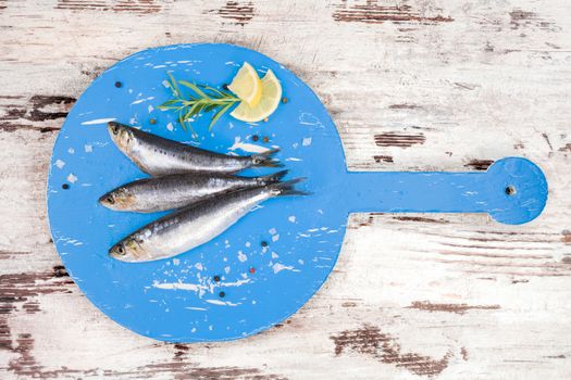 Three fresh anchovy fish on round blue wooden kitchen board on white and brown wooden table, top view. Culinary seafood concept. Delicious healthy eating.