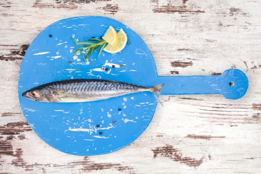 Delicious fresh mackerel fish on wooden kitchen board with lemon, rosemary and colorful peppercorns on white textured wooden background. Culinary healthy cooking.