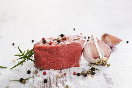 Big raw steak with peppercorns, garlic and fresh rosemary herbs on white wooden background. Culinary steak eating.