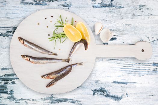 Delicious fresh sardines on wooden kitchen board with lemon, rosemary and colorful peppercorns on white textured wooden background. Culinary healthy cooking.