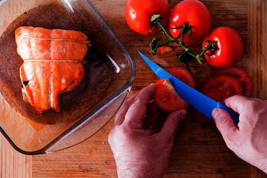 Close up view from above of the hands of a male chef slicing tomatoes with a blue knife to accompany an oven-baked salmon steak fresh from the oven on a wooden counter