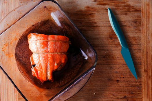 Fresh oven-baked salmon fresh from the oven in a glass oven dish alongside a sharp blue kitchen knife on a wooden kitchen counter ready to be served, view from above