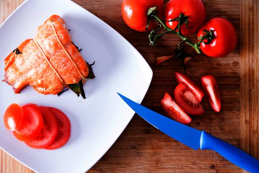 Delicious freshly roasted fresh salmon fillet served with fresh sliced vine tomatoes being prepared in a kitchen with a modern blue kitchen knife, close up view from above on a wooden counter