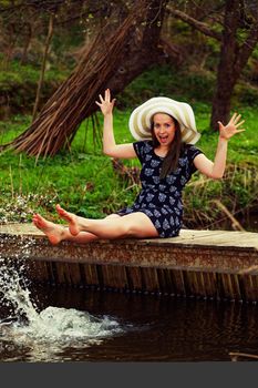 Cheerful fashionable woman sits on small bridge and splashing water by her legs in warm spring day.