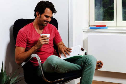 Young Man in Casual Clothing Sitting on Black Chair While Reading a Book and Holding a Glass of Drink.