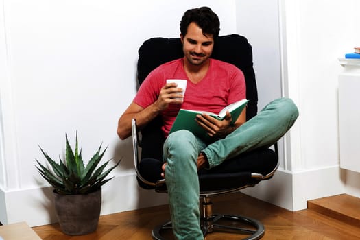 Young Man in Casual Clothing Sitting on Black Chair While Reading a Book and Holding a Glass of Drink.