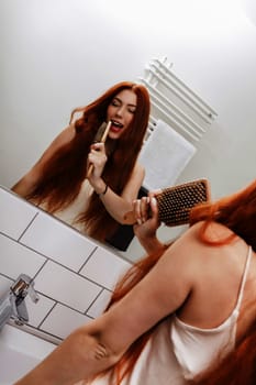 Close up Pretty Young Woman Brushing her Long Blond Hair In Front a Large Mirror in the Room.