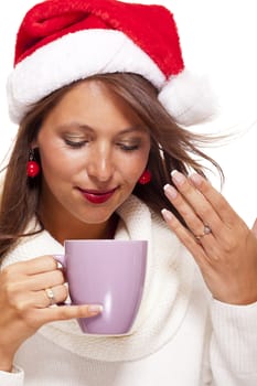 Cold attractive young woman with a cute smile in a festive red Santa hat sipping a hot mug of coffee that she is cradling in her hands to warm up in the winter weather, on white