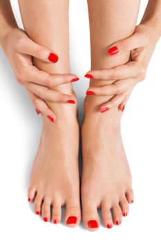 Woman with beautiful neatly manicured red finger and toenails sitting with bare feet clasping her ankles to display her nails, closeup on white in a fashion and beauty concept