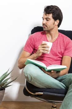 Young Man in Casual Clothing Sitting on Black Chair While Reading a Book and Holding a Glass of Drink.