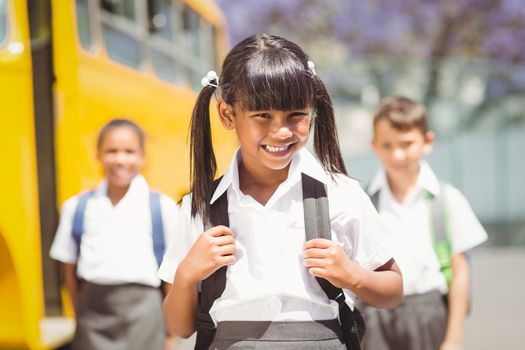 Cute pupil smiling at camera by the school bus outside the elementary school