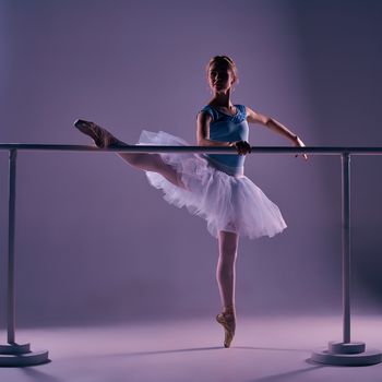 classic ballet dancer in white tutu posing on one leg at ballet barre on a lilac background