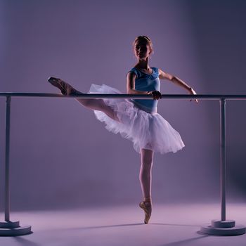 classic ballet dancer in white tutu posing on one leg at ballet barre on a lilac background