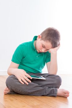 Close up Serious Male Kid Sitting on the Floor with Legs Crossed, Reading at his Tablet Computer While Leaning on his Hand.
