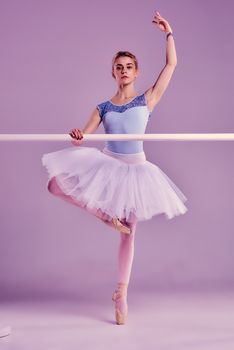 classic ballet dancer in white tutu posing on one leg at ballet barre on a lilac background