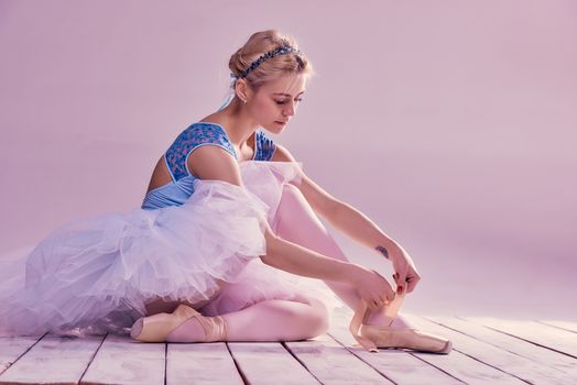 Professional ballerina putting on her ballet shoes on the wooden floor on a pink background
