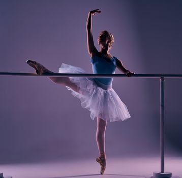 classic ballet dancer in white tutu posing on one leg at ballet barre on a lilac background