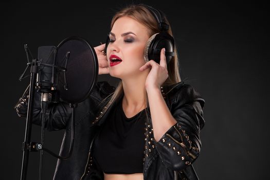 Portrait of a beautiful blonde young woman singing into microphone with headphones in studio on black background