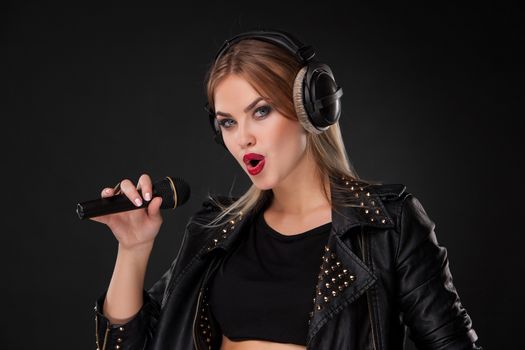 Portrait of a beautiful blonde young woman singing into microphone with headphones in studio on black background