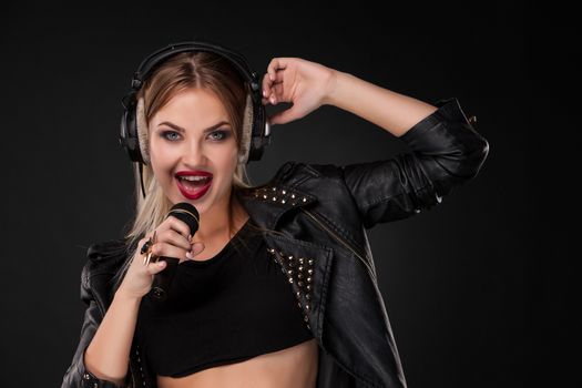 Portrait of a beautiful blonde young woman singing into microphone with headphones in studio on black background