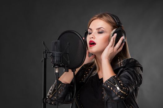 Portrait of a beautiful blonde young woman singing into microphone with headphones in studio on black background