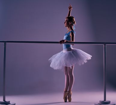 classic ballet dancer in white tutu posing at ballet barre on a lilac background